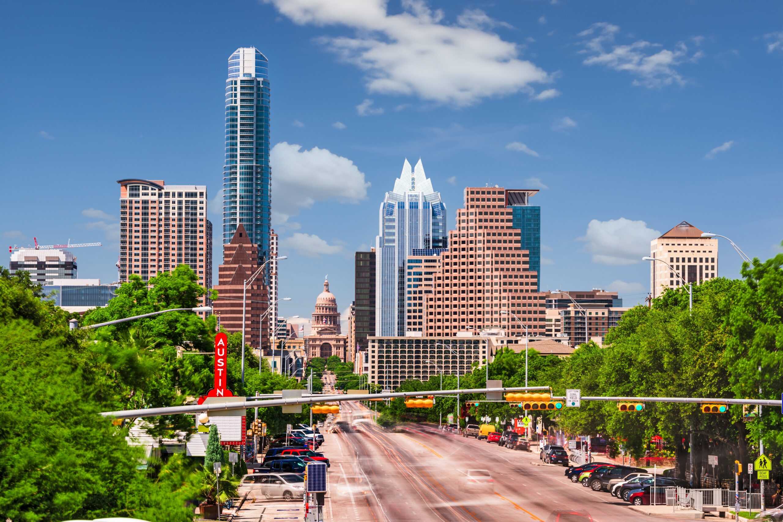 Austin, Texas skyline featuring the Texas State Capitol surrounded by modern skyscrapers, with a clear blue sky and light traffic on the main road.